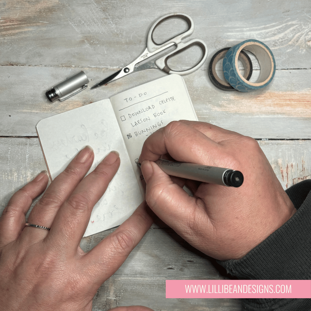 Person writing in a small notebook with stationery items on a wooden surface