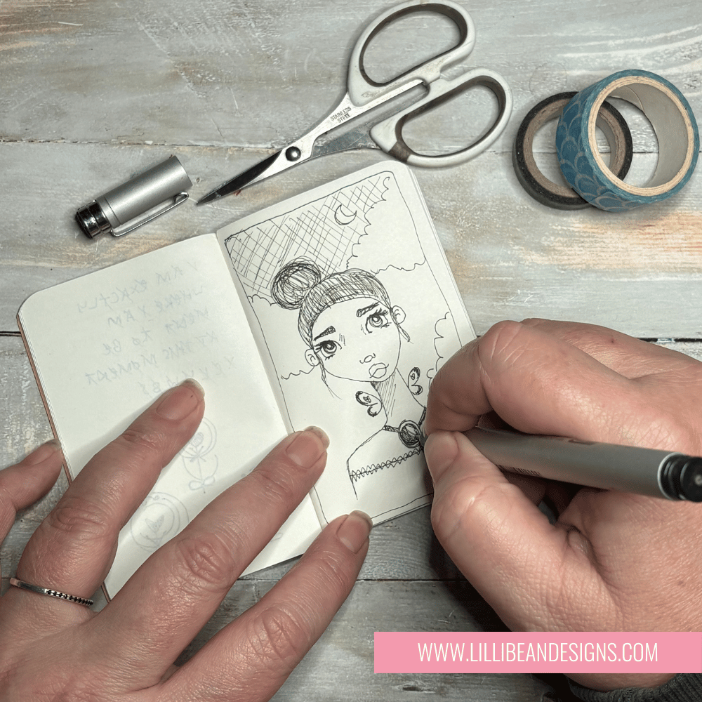 Person drawing a character in a notebook with stationery items on a wooden surface.