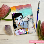 Artistic journal with a girl and lamb illustration on a wooden surface with flowers and tools.