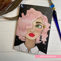 Illustration of a woman with pink hair and red lipstick on a black background, with a pen and plant in the background. GREETING CARD