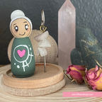 Small figurine with a green shirt featuring a pink heart on a wooden stand with crystals and flowers in the background.