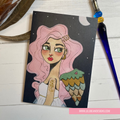 Illustration of a woman with pink hair and winged shoulder on a black background, with a pen and feather in the background. GREETING CARD