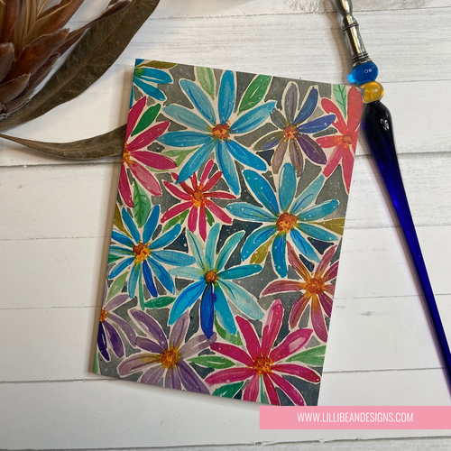 Colorful floral design on a small card with a paintbrush and plant on a white surface.
