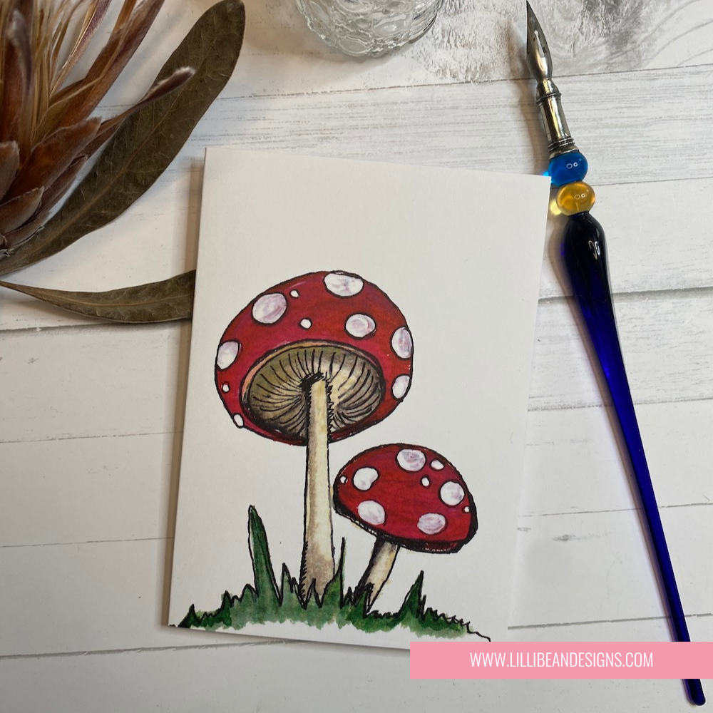 Illustration of red mushrooms with white spots on a white background, with a paintbrush and glass container in the background. GREETING CARD