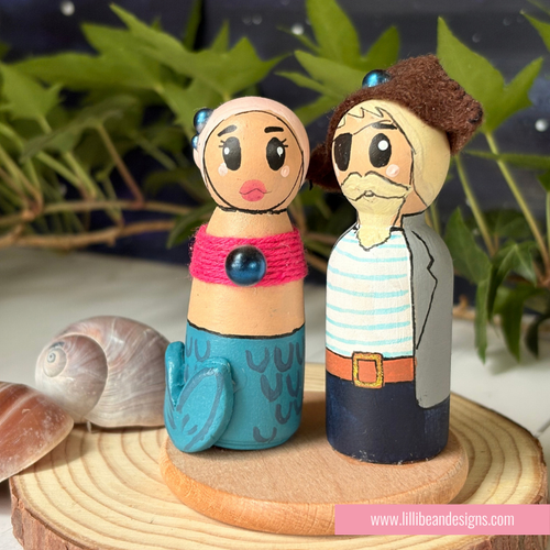 Handcrafted wooden peg dolls representing a mermaid and a sailor, displayed on a wooden slice with seashells and green foliage in the background.