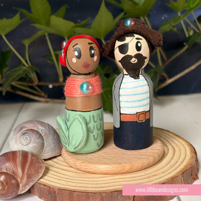 Handcrafted wooden peg dolls representing a mermaid and a pirate, displayed on a round wood slice with seashells and green leaves in the background.