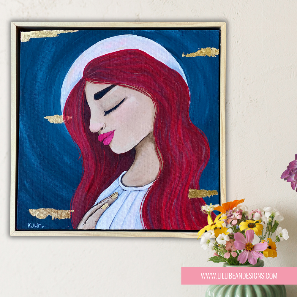 Framed artwork of a woman with red hair and a blue background, displayed on a wall with a small bouquet of flowers.