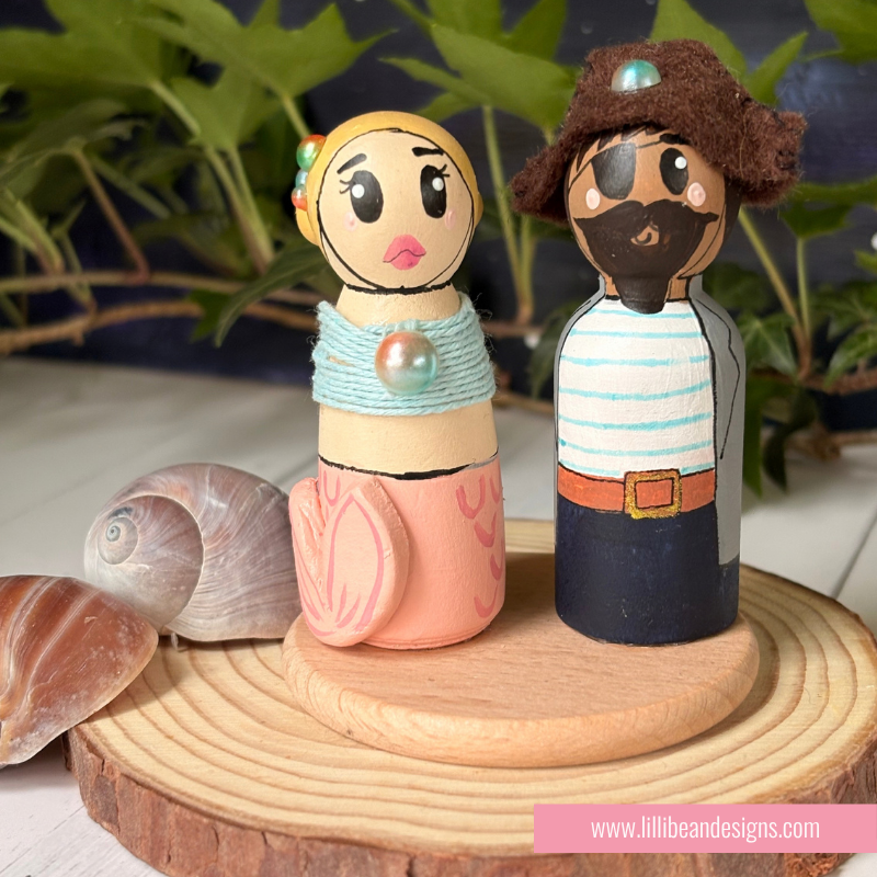 Hand-painted wooden peg dolls styled as a mermaid and a pirate, displayed on a round wooden slice with seashells and green foliage in the background.