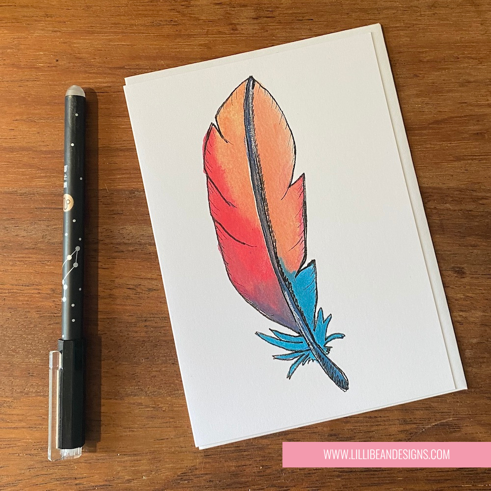 Card with a colorful feather design on a wooden surface with a pen next to it.