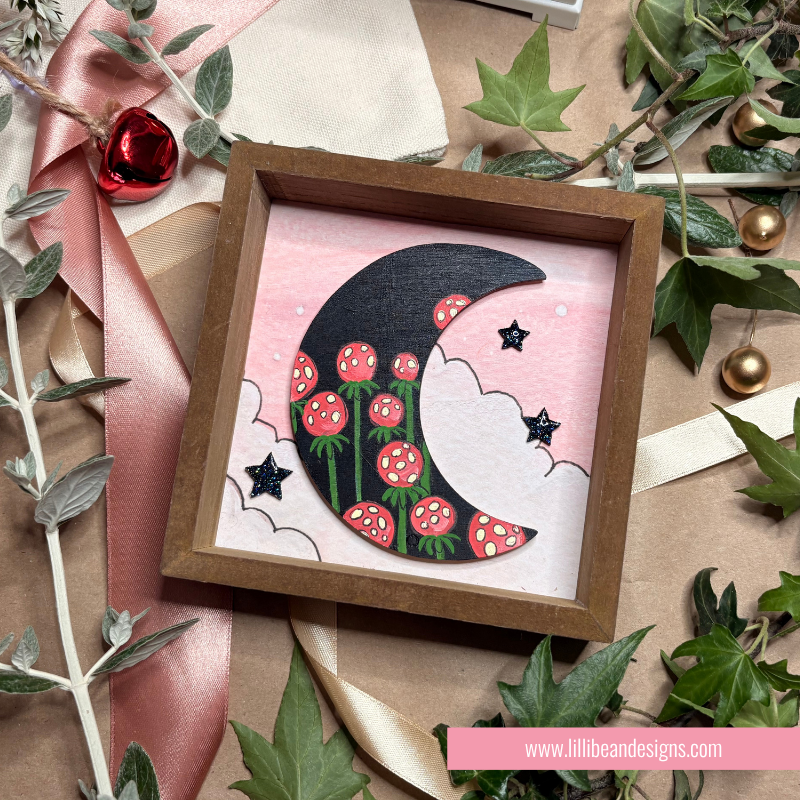 A square wooden shadow box containing a whimsical illustration of a black crescent moon with red and white flowers, green stems, and small black stars on a pink background. The box is styled on a kraft paper surface with ribbon, greenery, and small red and gold ornaments.