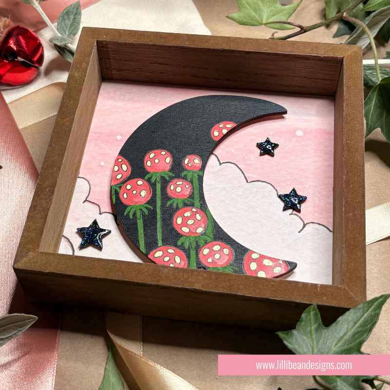 A square wooden shadow box display featuring a hand-painted black crescent moon decorated with red flowers and green stems, set against a pink background with white clouds and small black star embellishments.