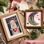 Two framed art prints displayed on a kraft paper surface with decorative greenery, ribbons, and small red ornaments. The left frame shows a stylized angel figure with wings holding a red heart that says 'HOLY', and the right frame shows a crescent moon with flowers and stars on a pink background.