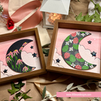 Two framed handmade paper moon and star art pieces displayed on a styled surface with ribbon, leaves, and a lantern. The left frame shows a black crescent moon with pink flowers, and the right frame shows a dark gray crescent moon with red strawberries, green leaves, and white flowers on a pink cloud background.