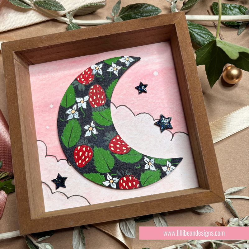 A square wooden shadow box frame containing a handmade paper craft of a crescent moon decorated with green leaves, red strawberries, and white flowers on a pink background with small black stars.