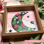 A wooden shadow box display featuring a crescent moon design decorated with green leaves, red strawberries, and small white flowers on a pink background with cloud outlines. The box is styled with decorative leaves and ribbon.