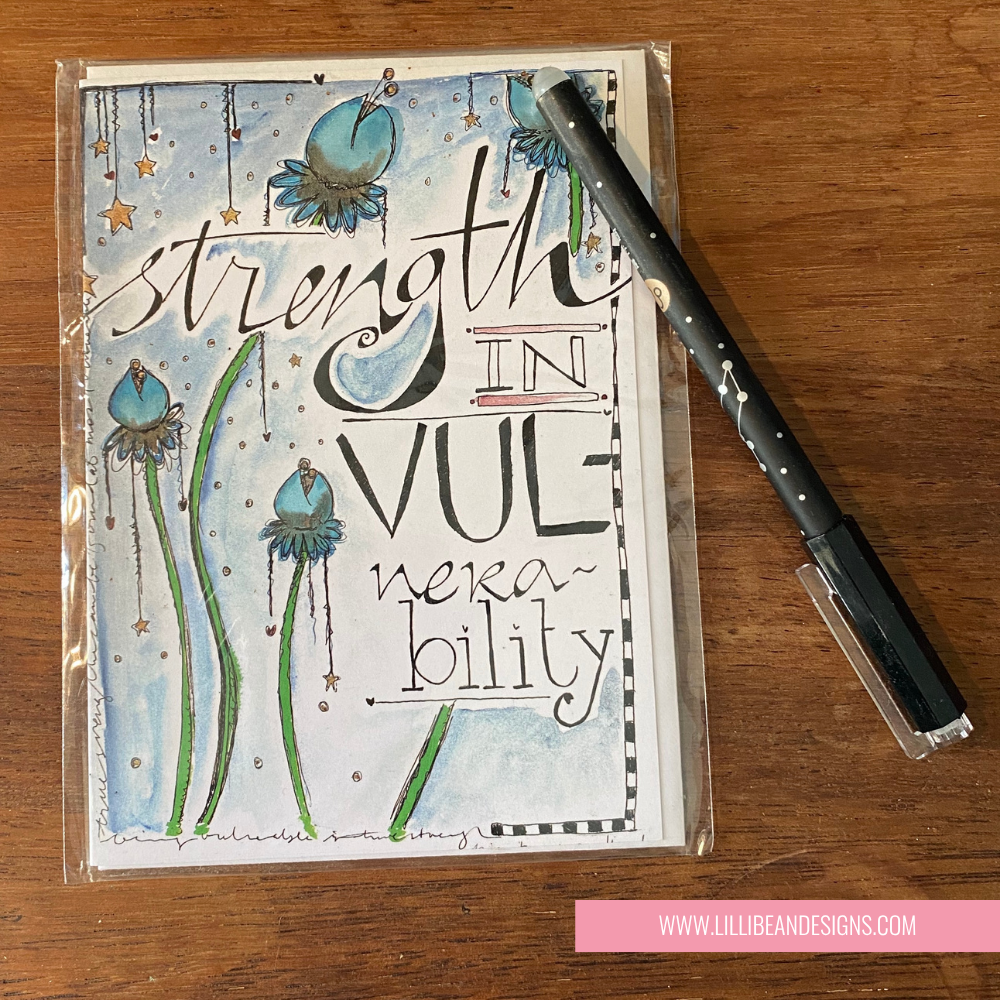 Handmade card with motivational quote 'Strength in Vulnerability' on a wooden surface with a pen.
