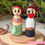 Two handcrafted wooden mermaid and pirate-themed dolls standing on a round wooden base, with a blurred green plant background and seashells nearby.