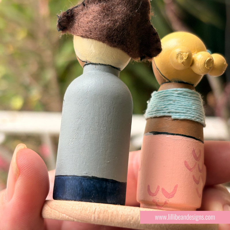 A close-up of a hand holding two handcrafted wooden peg dolls outdoors. One doll has a brown felt hat and a light blue painted body with a dark blue base.