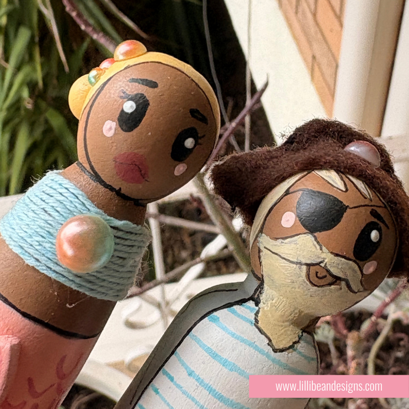 Two hand-painted wooden toy figures with painted faces, one wearing a yellow hat and light blue wrap, and the other wearing a brown hat and striped shirt, photographed outdoors with a pink watermark at the bottom.
