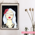 A framed art print featuring a stylized illustration of a blindfolded woman with white hair, red lips, and a red flower on her head, hanging on a white wall next to dried flowers in a glass vase.
