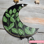 Decorative wall hanging with green leaf pattern on a black crescent moon shape, displayed on a wooden surface.