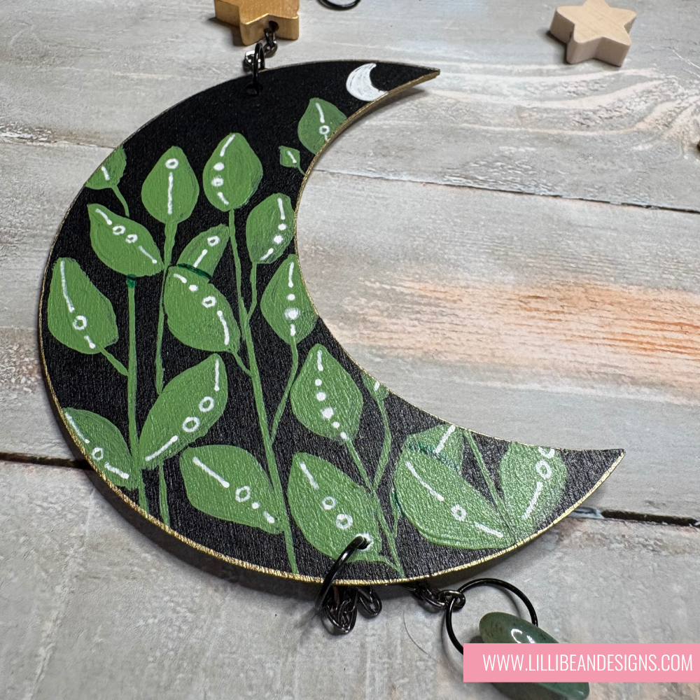 Decorative wall hanging with green leaf pattern on a black crescent moon shape, displayed on a wooden surface.