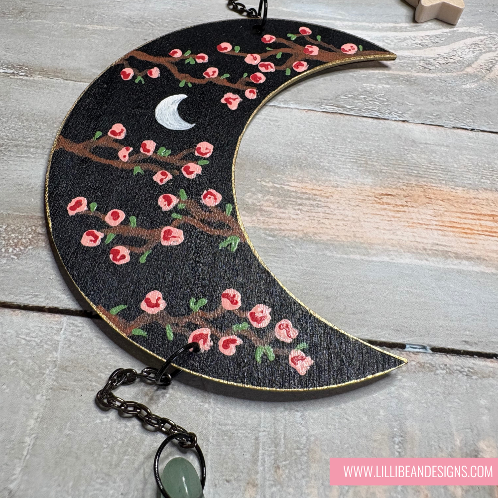 Decorative crescent moon with floral design and chain on a wooden surface, featuring LilliBean Designs branding.