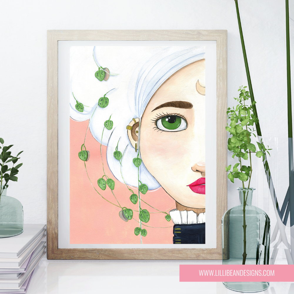 A framed art print featuring a stylized illustration of a woman with green eyes, white hair, and pink lips, with green trailing vines on a peach background. The print is in a light wood frame and styled on a white surface with decorative plants and books.