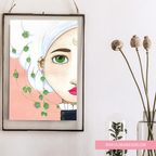 A framed art print featuring a stylized portrait of a woman with white hair, green eyes, and pink lips, hanging on a white wall next to dried flowers in a glass vase. The print has a pink background and green leaf motifs.