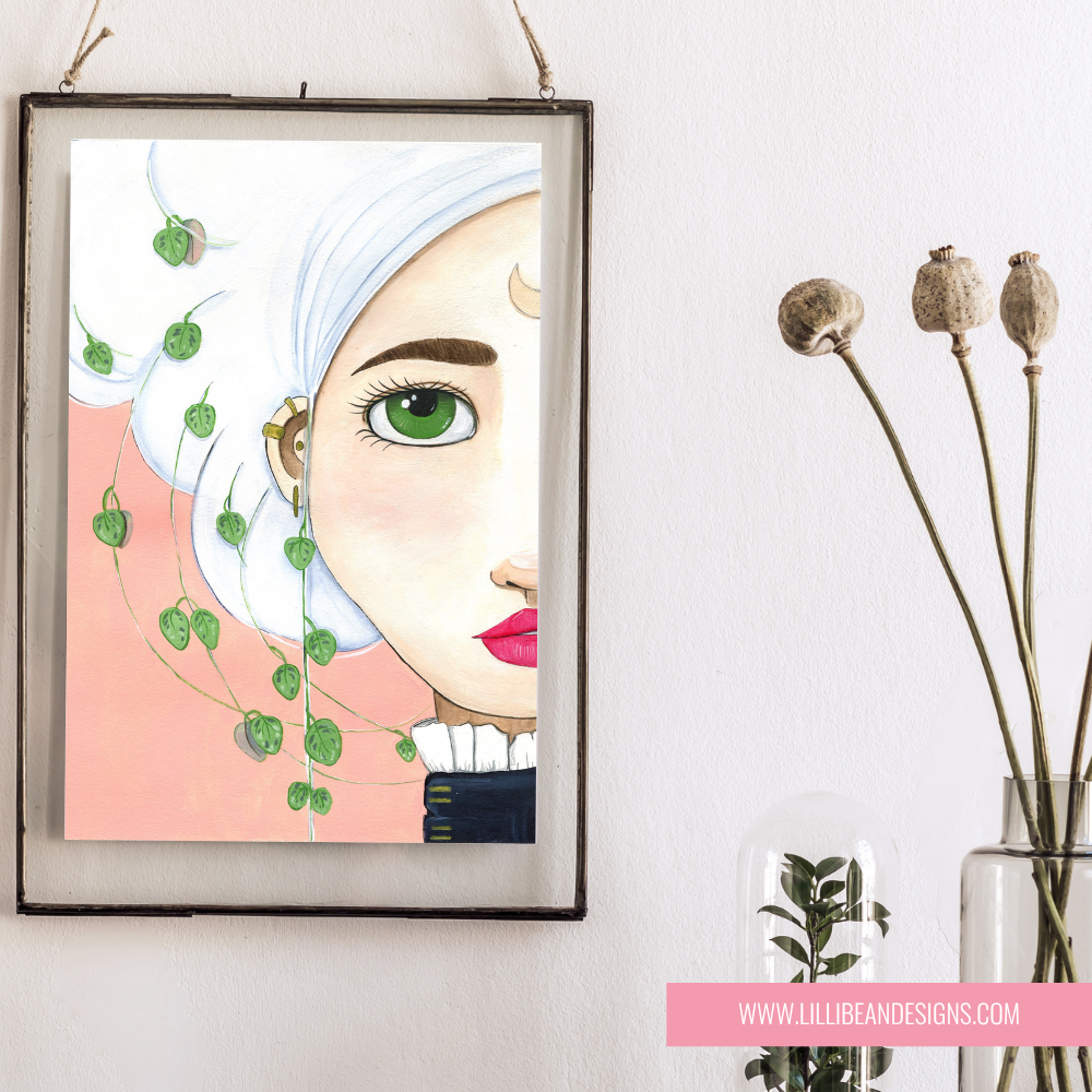 A framed art print featuring a stylized portrait of a woman with white hair, green eyes, and pink lips, hanging on a white wall next to dried flowers in a glass vase. The print has a pink background and green leaf motifs.