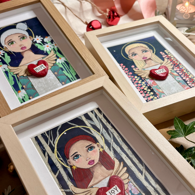 Three framed art prints featuring stylized female figures with angel wings and hearts labeled 'JOY' and 'LOVE', arranged on a tabletop with warm decorative props.