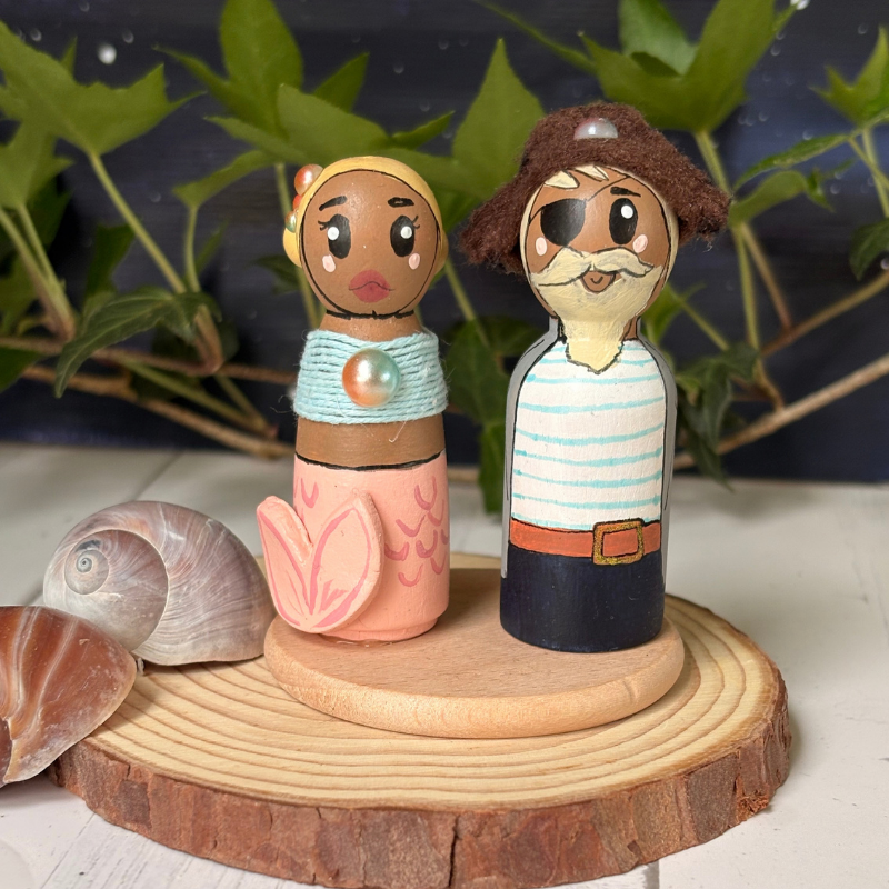 Hand-painted wooden couple figurines styled on a natural wood slice with seashells and green leaves in the background.