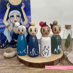 Set of four wooden peg dolls with unique designs on a wooden stand, with a painting and small bottle in the background.