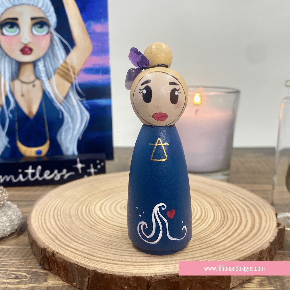 Wooden peg doll with blue body and white design on a wooden surface with a candle in the background.
