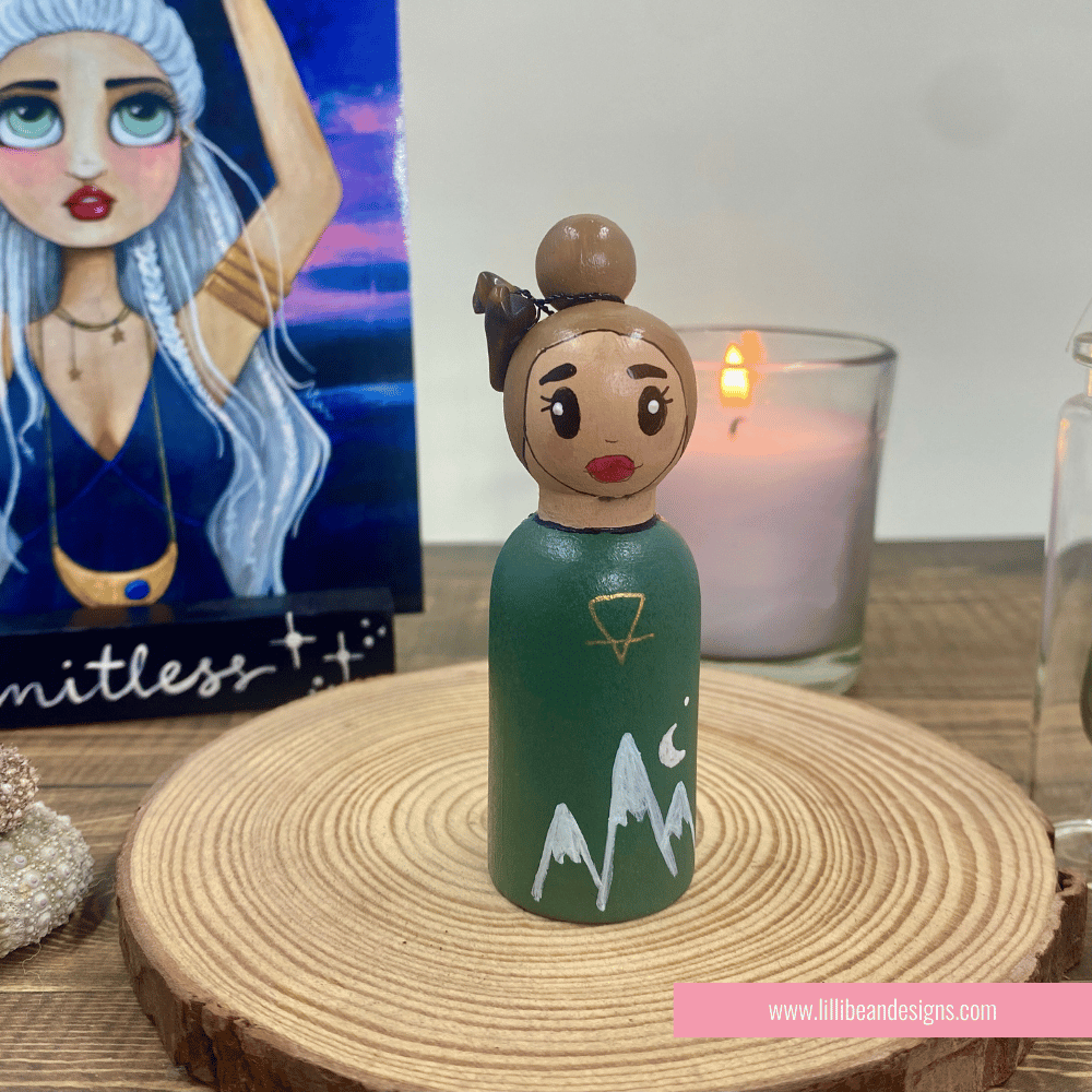 Small figurine with green dress and mountain design on a wooden surface with a candle in the background.