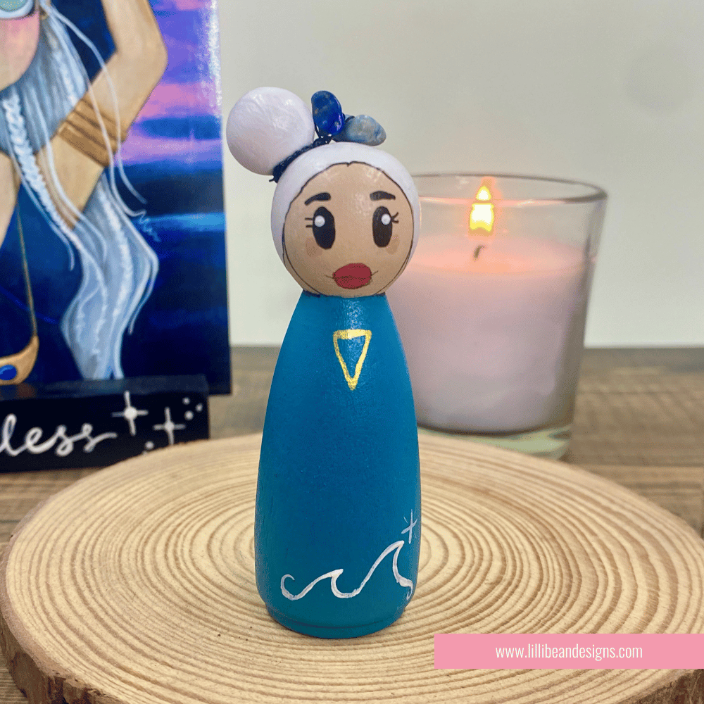Blue peg doll with white hair and red lips on a woven mat with a candle in the background.