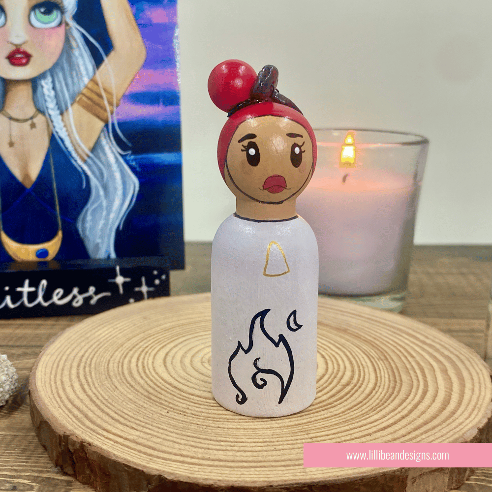 Peg doll with red hair and white dress on a wooden surface with a candle in the background.