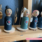 Set of peg dolls with earrings on a wooden stand, featuring a brand logo at the bottom.
