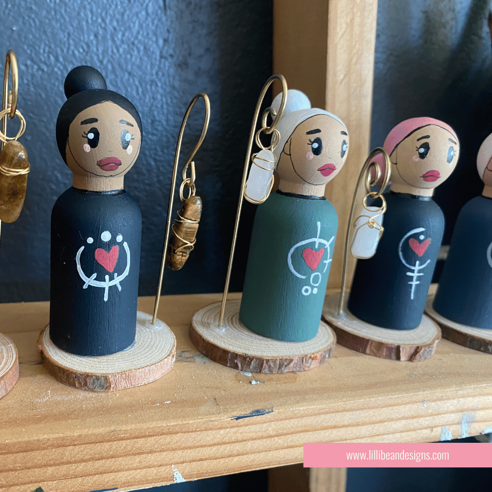 Set of peg dolls with earrings on a wooden stand, featuring a brand logo at the bottom.
