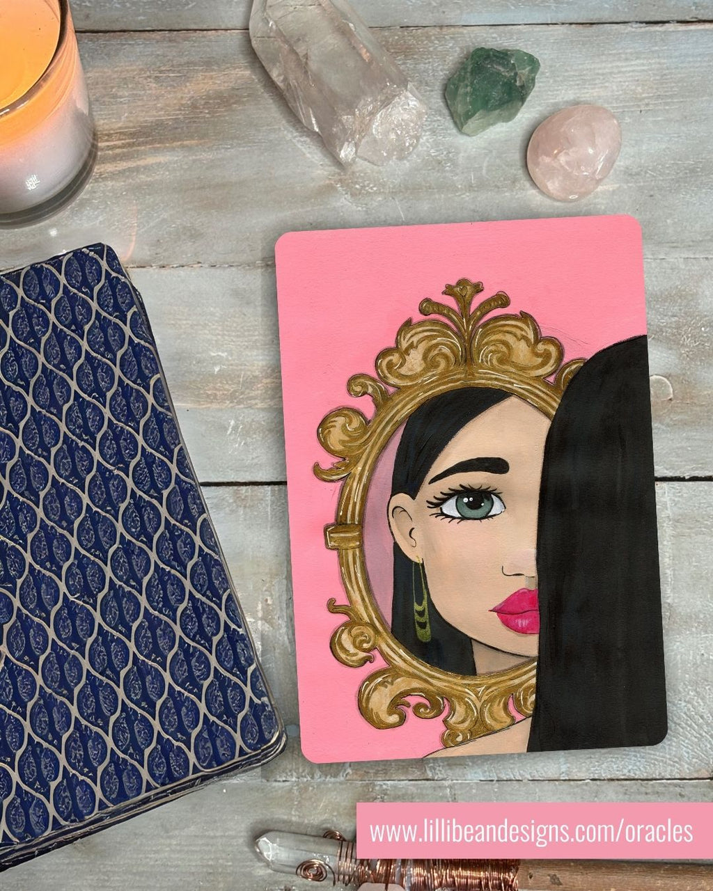 Card with a stylized portrait of a woman in front of a mirror on a pink background, placed on a wooden surface with crystals and a candle.