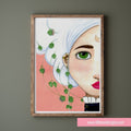 Framed artwork of a woman's face with green leaves on a pink background, displayed on a wall.
