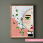 Framed artwork of a woman's face with green leaves on a pink background, displayed on a wall.