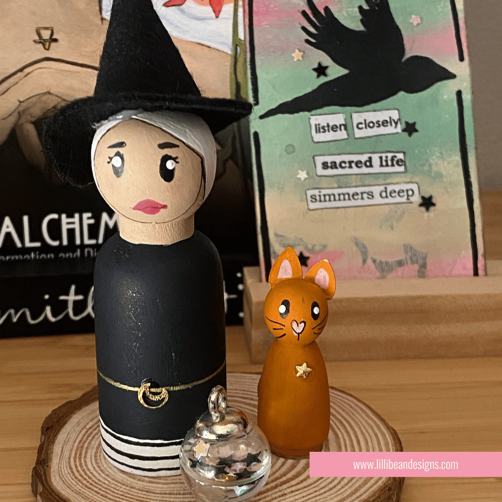 Wooden witch figurine with a cat on a wooden base, with a blurred background featuring books and stationery items. LilliBean Designs