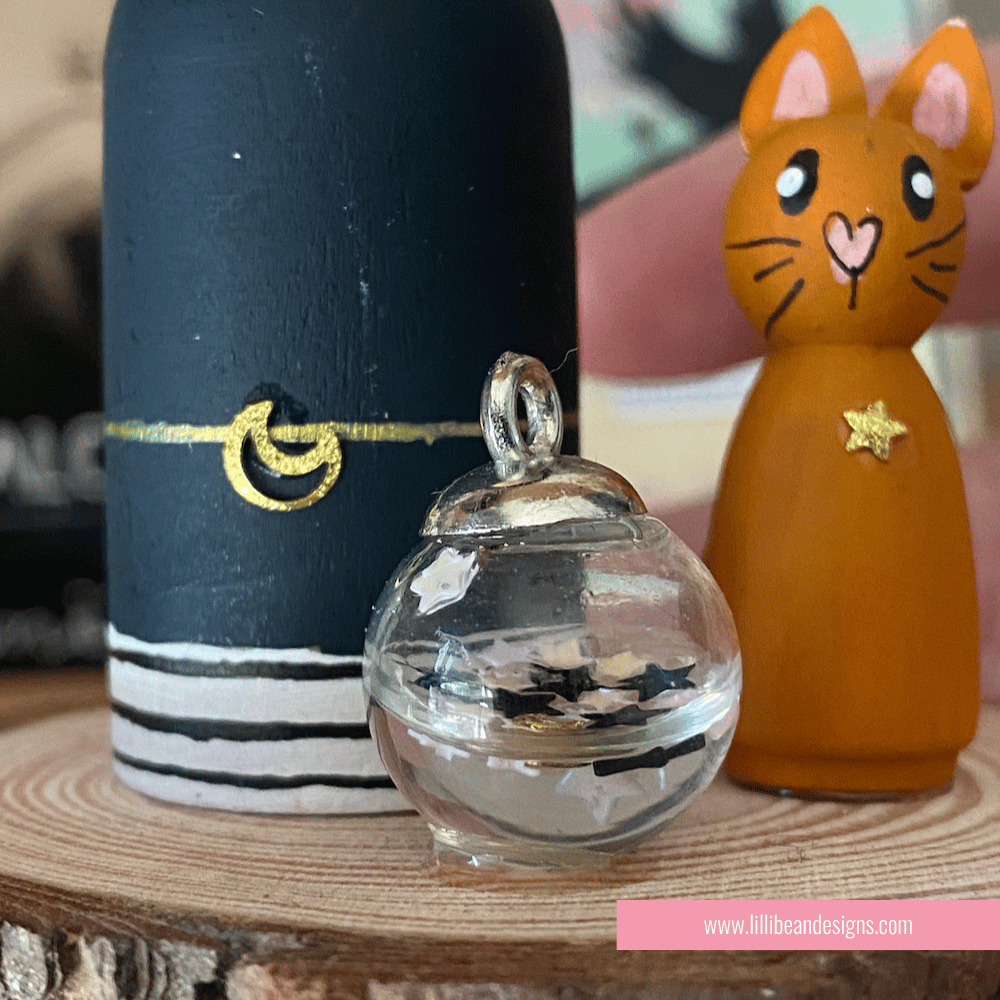 Wooden witch figurine with a cat on a wooden base, with a blurred background featuring books and stationery items. LilliBean Designs