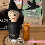 Wooden peg doll of a witch with a cat and small jar on a wooden surface, with 'LillieBeads Designs' branding.