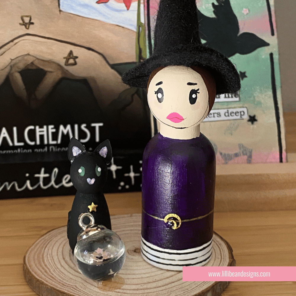 Wooden witch figurine with a cat on a wooden base, with a blurred background featuring books and stationery items. LilliBean Designs