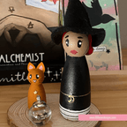 Wooden peg doll of a witch with a cat and lantern on a wooden surface, with 'Alchemist' and 'Mithli' branding in the background.