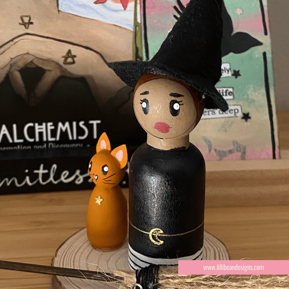 Wooden witch figurine with a cat on a wooden base, with a blurred background featuring books and stationery items. LilliBean Designs