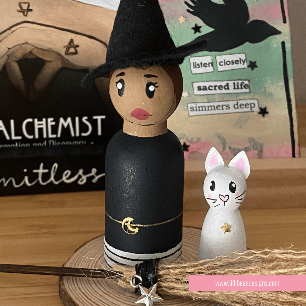 Wooden witch figurine with a cat on a wooden base, with a blurred background featuring books and stationery items. LilliBean Designs